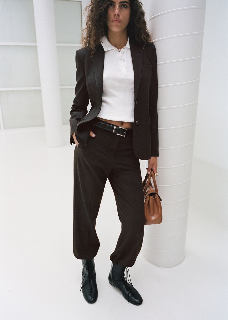 Woman in a brown blazer and trousers stands in a white studio, carrying a tan purse. Wearing black lace-up boots, with curly hair.