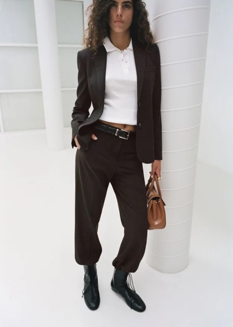 Woman in a brown blazer and trousers stands in a white studio, carrying a tan purse. Wearing black lace-up boots, with curly hair.