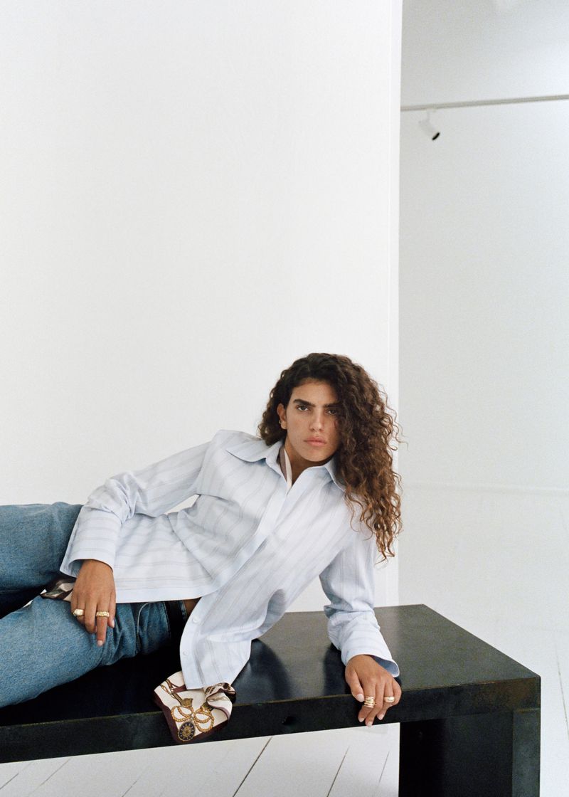 Young adult woman with curly brown hair, wearing a striped shirt and jeans, lounges on a black table in a white room.