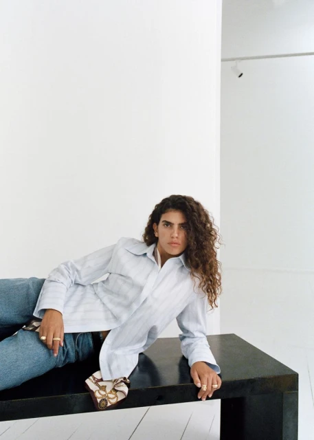 Young adult woman with curly brown hair, wearing a striped shirt and jeans, lounges on a black table in a white room.