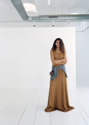 Model wearing a long brown dress with a sweater tied around her waist in a white studio space