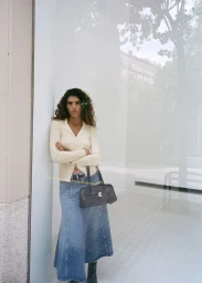 Model Sun Mizrahi leaning against a wall wearing a cream cardigan and denim skirt for Mango