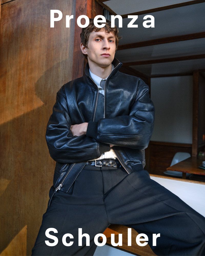 A young adult male model wearing a black leather bomber jacket and black trousers poses with crossed arms in a fashion editorial studio shot.