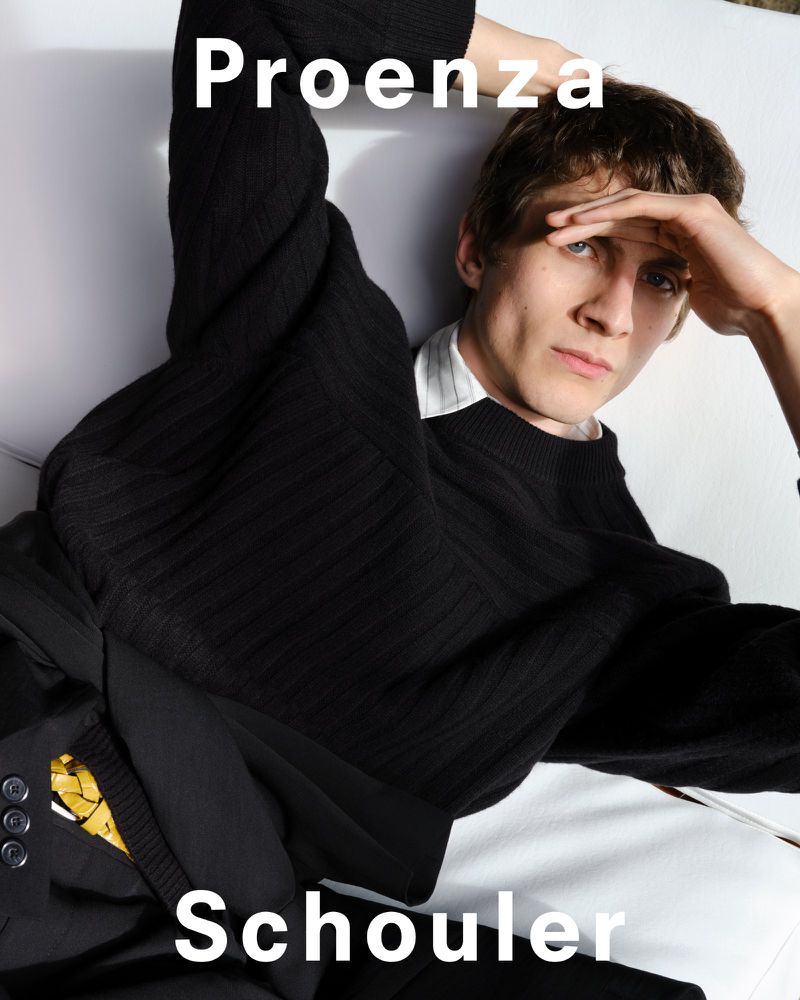 A light-skinned young man is lying down wearing a black sweater with visible clothing brand names, Proenza Schouler, in the image.