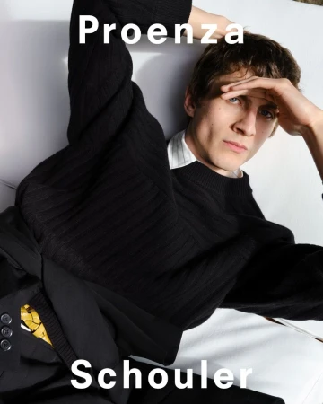 A light-skinned young man is lying down wearing a black sweater with visible clothing brand names, Proenza Schouler, in the image.