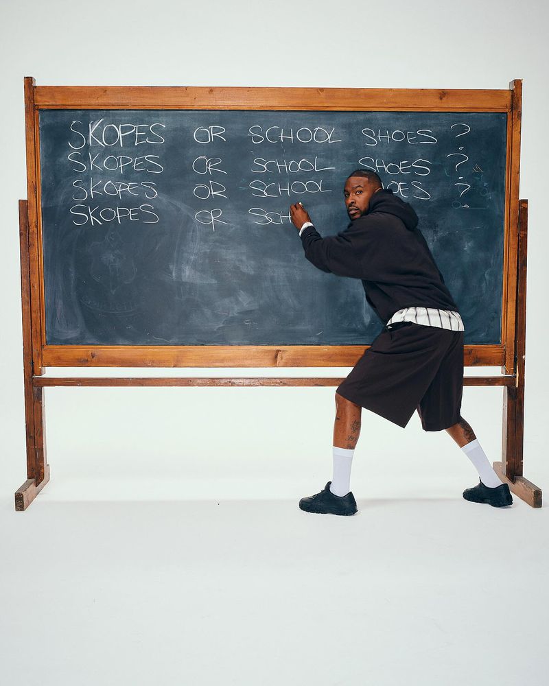 Adult male writing 'Skopes or School Shoes?' on a blackboard in a studio. He wears a dark hoodie, shorts, socks, and shoes, facing the camera.