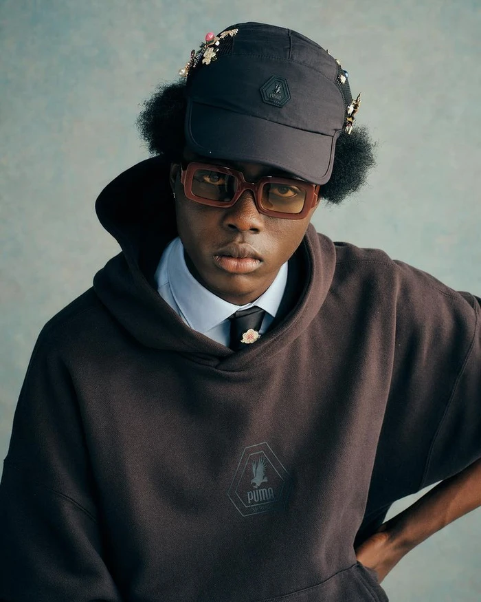 Portrait of a man wearing a dark cap, glasses, and a Puma hoodie against a textured grey background.