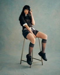 Model seated on a metal stool wearing a dark short-sleeve top, shorts, and black sneakers against a mottled blue backdrop.