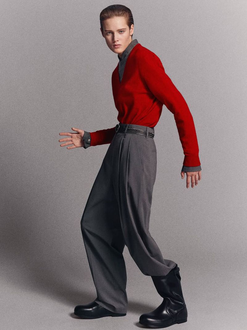 A person in a red sweater and gray pants strikes a pose in a studio setting. The person is wearing black boots.