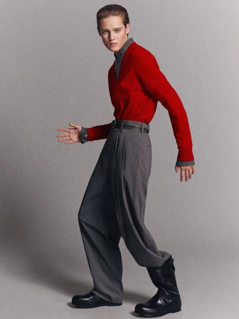A person in a red sweater and gray pants strikes a pose in a studio setting. The person is wearing black boots.