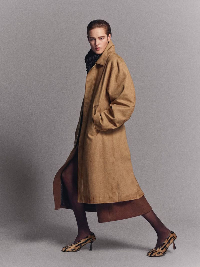 Fashionable woman in stylish tan coat and animal print heels poses in a photo studio, showcasing an elegant and modern look.