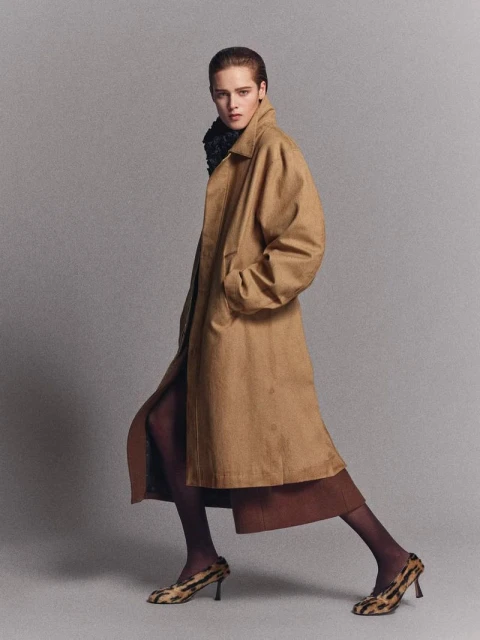 Fashionable woman in stylish tan coat and animal print heels poses in a photo studio, showcasing an elegant and modern look.