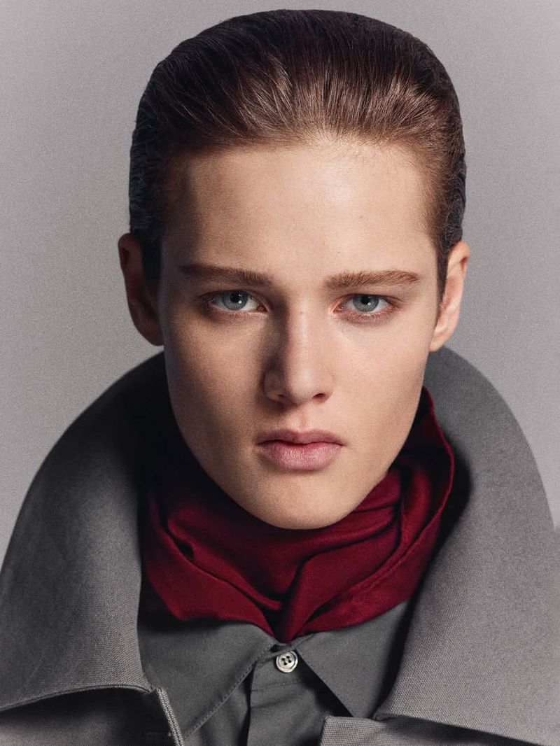 Portrait of a young adult wearing a gray shirt and coat with a maroon scarf, set against a muted gray background in a photo studio.