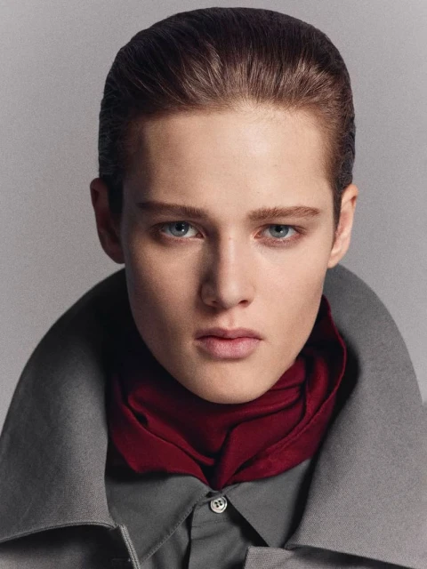 Portrait of a young adult wearing a gray shirt and coat with a maroon scarf, set against a muted gray background in a photo studio.