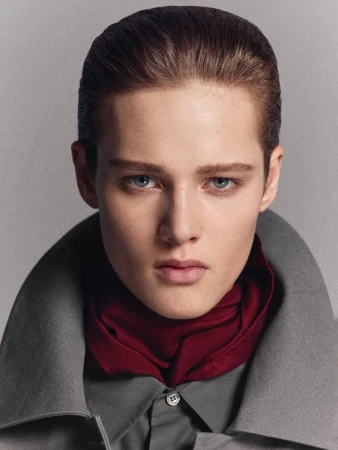 Portrait of a young adult wearing a gray shirt and coat with a maroon scarf, set against a muted gray background in a photo studio.