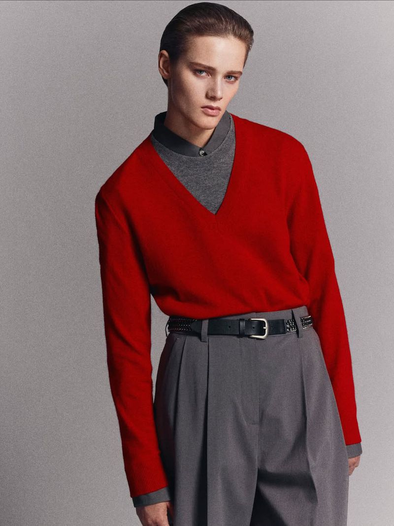 Fashion shot of a person with short hair wearing a red sweater and gray trousers against a neutral gray backdrop in a studio setting.