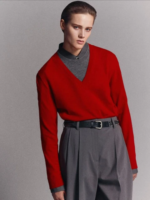 Fashion shot of a person with short hair wearing a red sweater and gray trousers against a neutral gray backdrop in a studio setting.