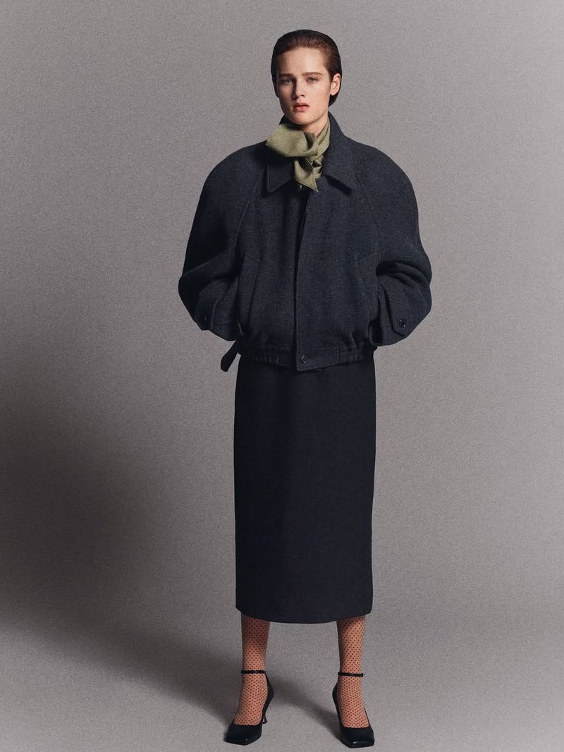 Elegant woman poses in a navy jacket, midi skirt, and black heels against a neutral grey backdrop, embodying classic fashion.