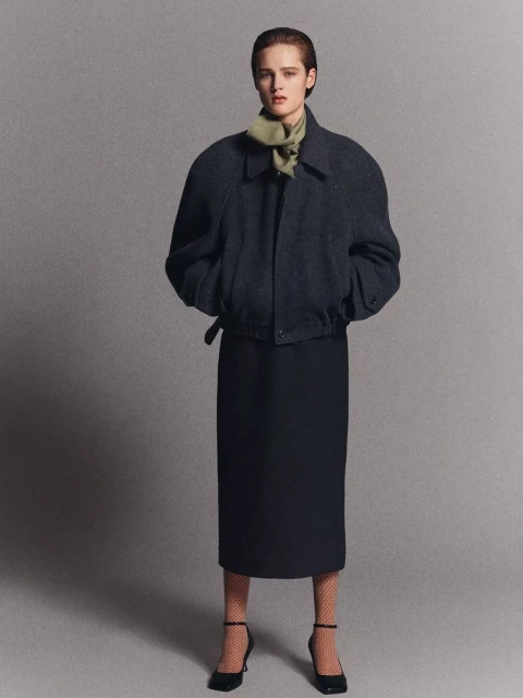 Elegant woman poses in a navy jacket, midi skirt, and black heels against a neutral grey backdrop, embodying classic fashion.