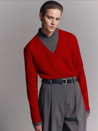 Model wearing a bright red v-neck sweater over a grey shirt and pleated trousers against a neutral grey studio background.