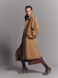 Model wearing a tan coat and patterned heels posing against a grey seamless studio backdrop.