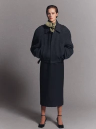 Model wearing a dark wool jacket and matching skirt with an olive neck scarf against a neutral studio backdrop.