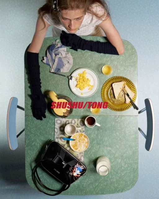 Overhead shot of a young woman in a white dress, sitting at a green table with breakfast foods, a handbag, and morning light.