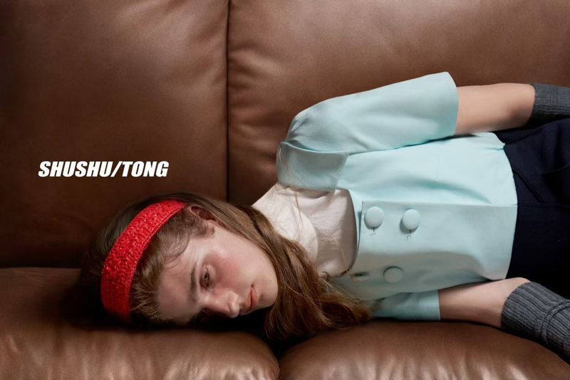 A young adult female model rests on a brown leather sofa with a light blue jacket and a red headband for a fashion advertisement.