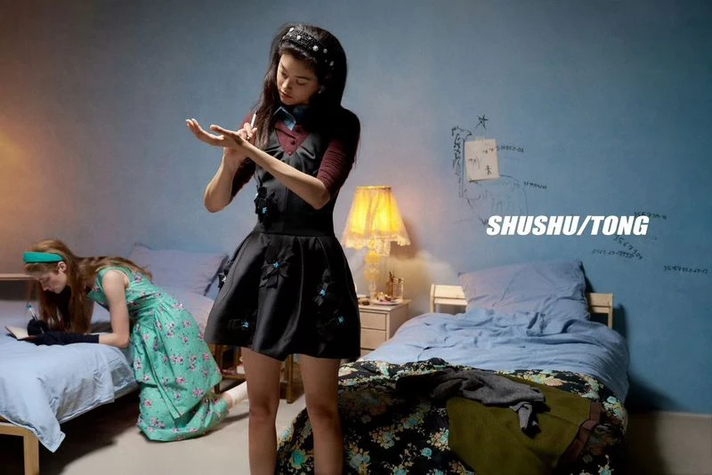 Two young women pose in a vintage-style bedroom; one stands applying makeup while the other kneels on a nearby bed reading a book.