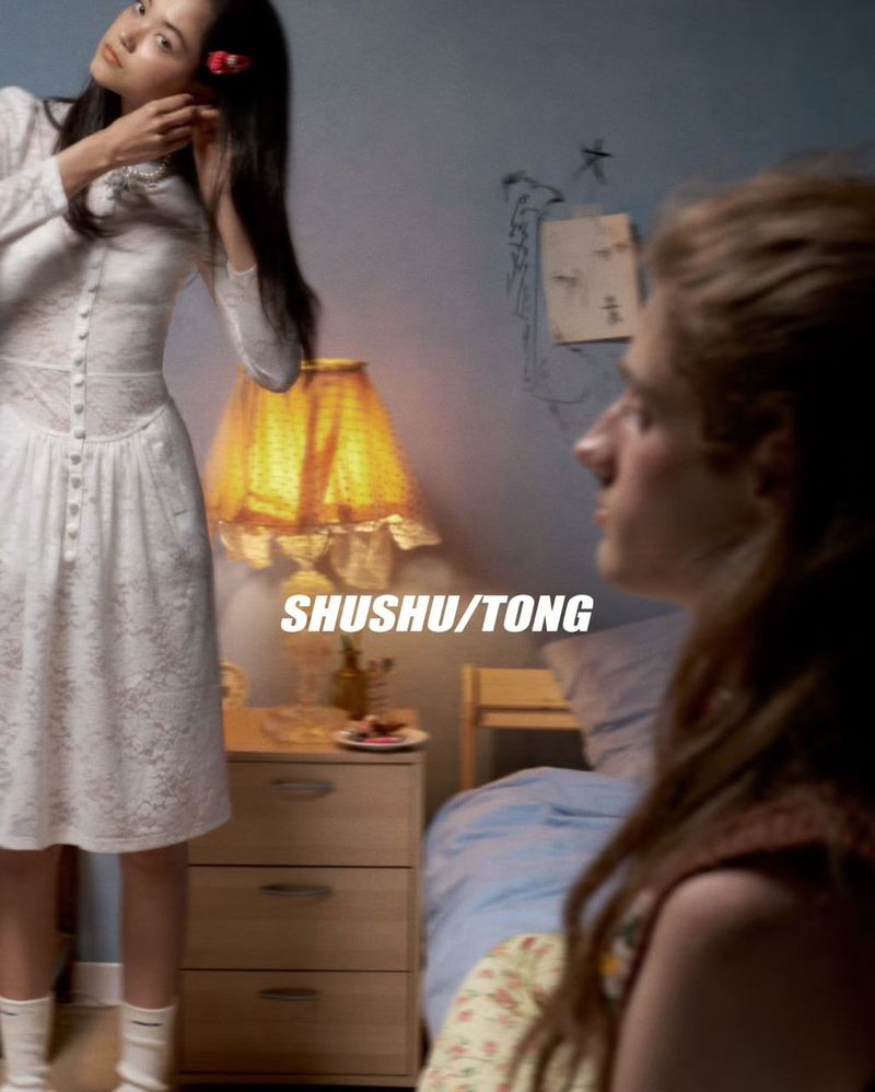 Two young women indoors, one wearing a white lace dress, standing near a nightstand with a lamp, with one figure partially blurred in the foreground.