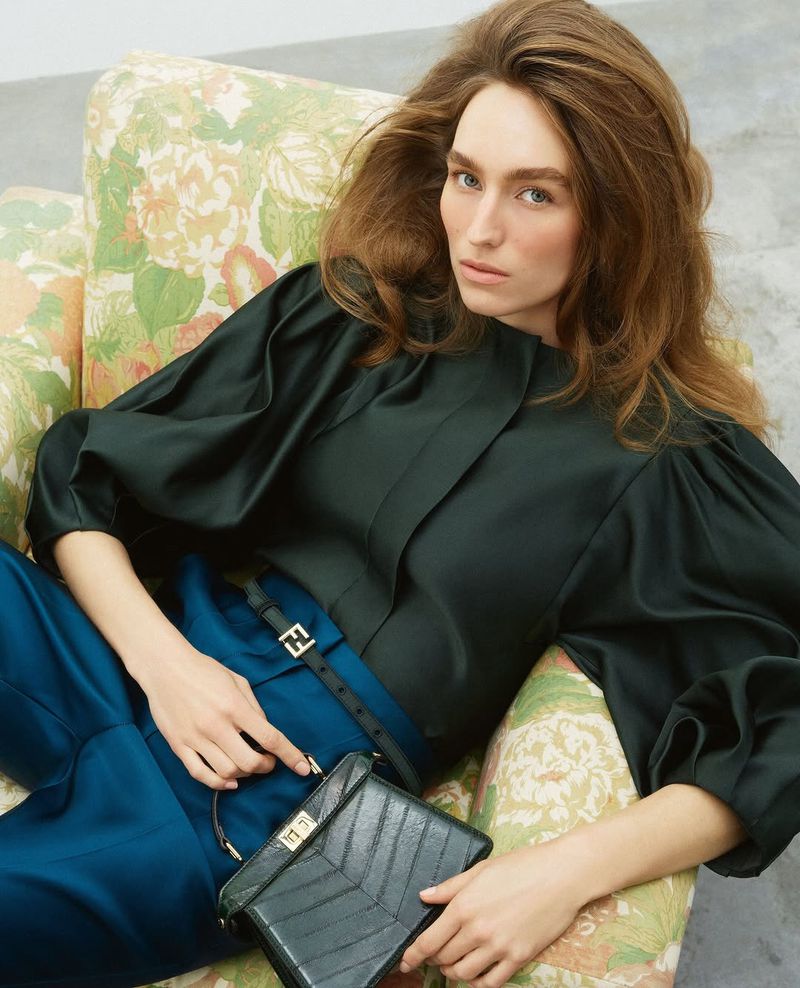 Elegant woman in a green blouse and blue pants sits on a floral couch, holding a black purse in a brightly lit indoor setting.
