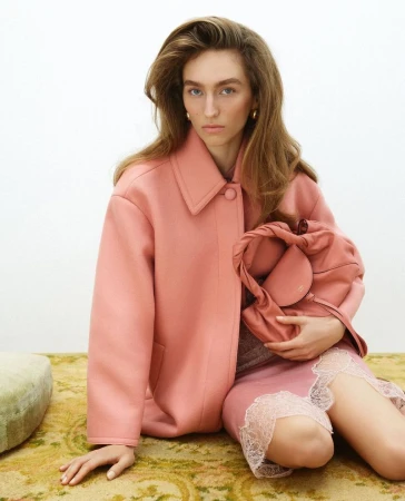 A woman in a pink coat poses while seated and holding a pink purse in a studio environment, creating a fashion-forward and minimalist aesthetic.