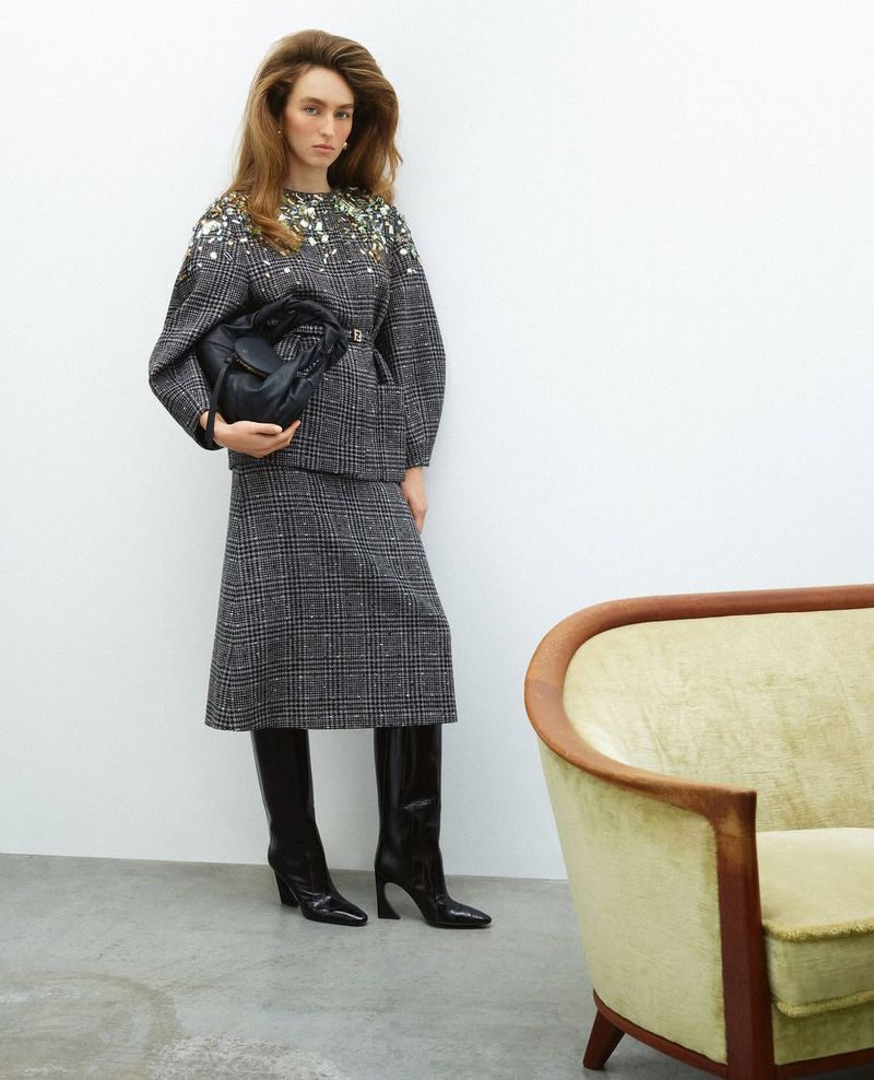 A woman poses in a grey plaid skirt suit and black boots, holding a black bag, in a fashion studio setting with a cream chair.