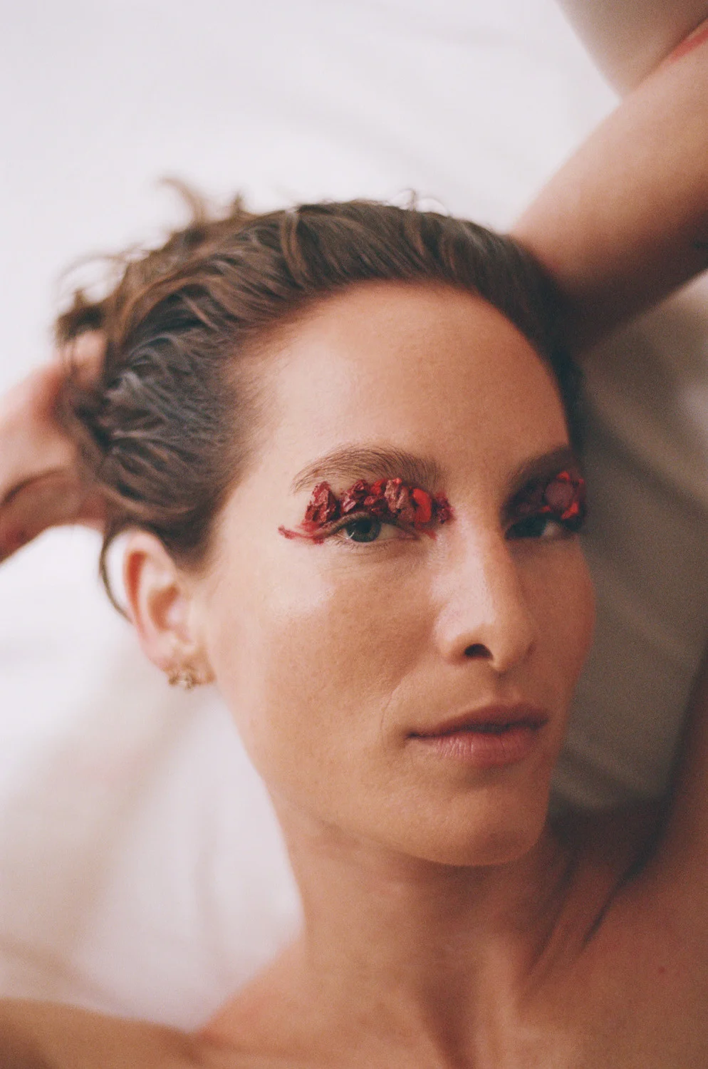 Close-up portrait of a woman with floral eye makeup, resting with hands behind her head against a white background, soft studio lighting.