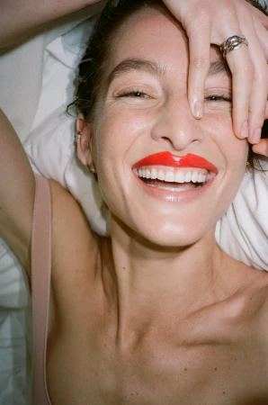 Close-up portrait of a smiling woman with vibrant red lipstick, hand raised to her forehead against white bedding