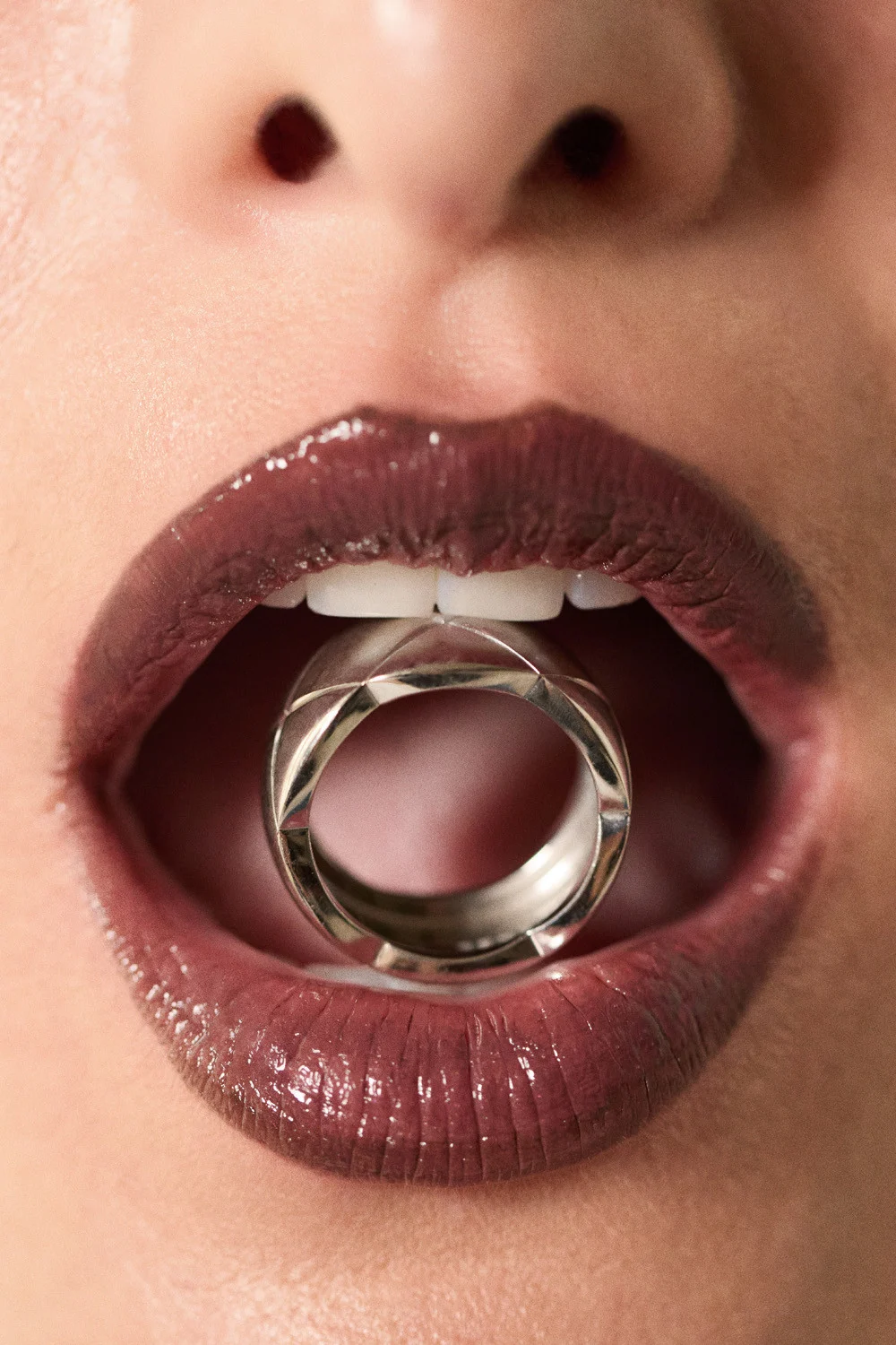Close-up macro shot of dark glossy lips holding a metallic silver ring between teeth for Love Want jewelry editorial