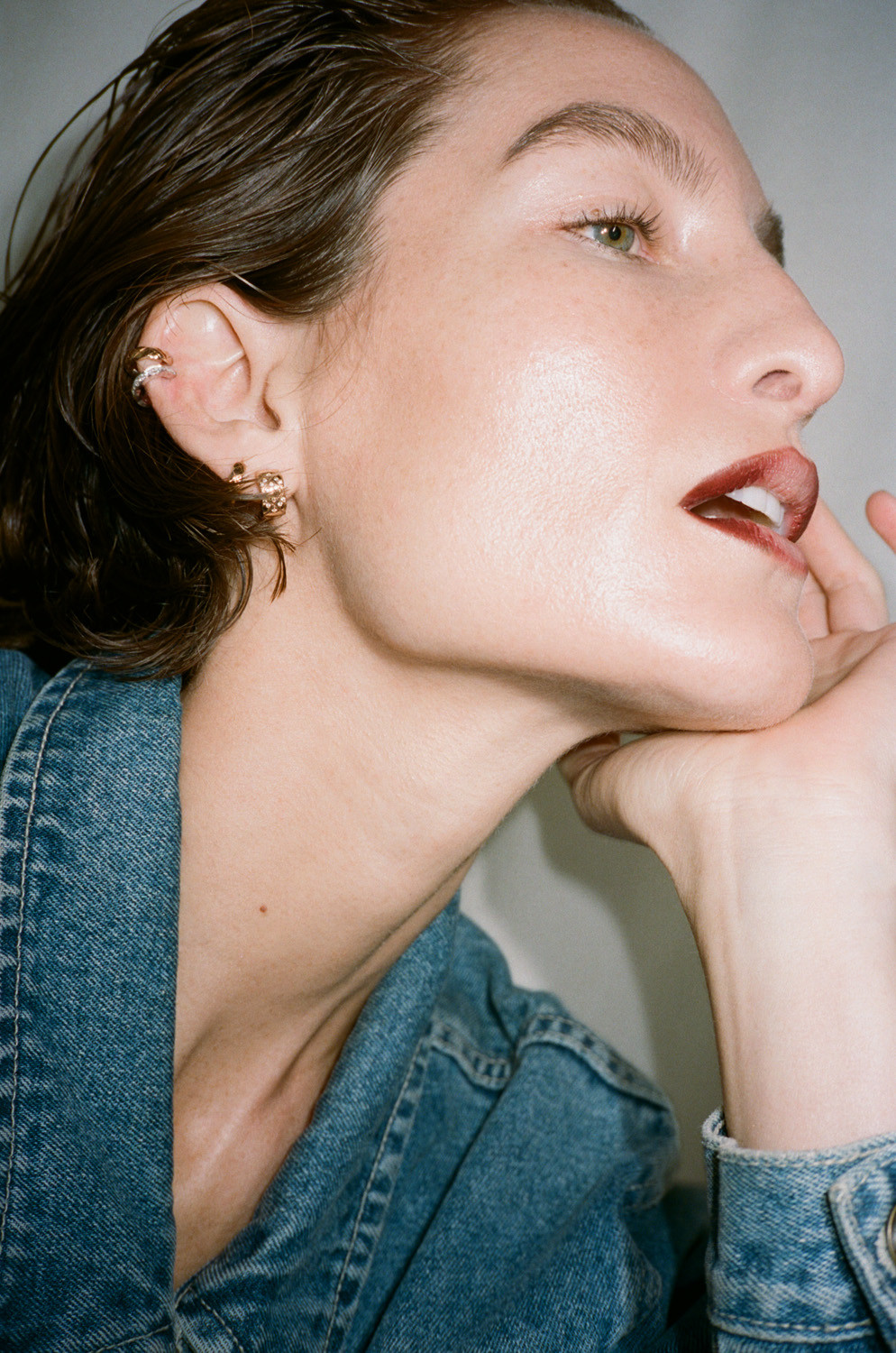 Close-up profile of a woman with wet hair wearing a denim shirt and gold earrings, resting her chin on her hand