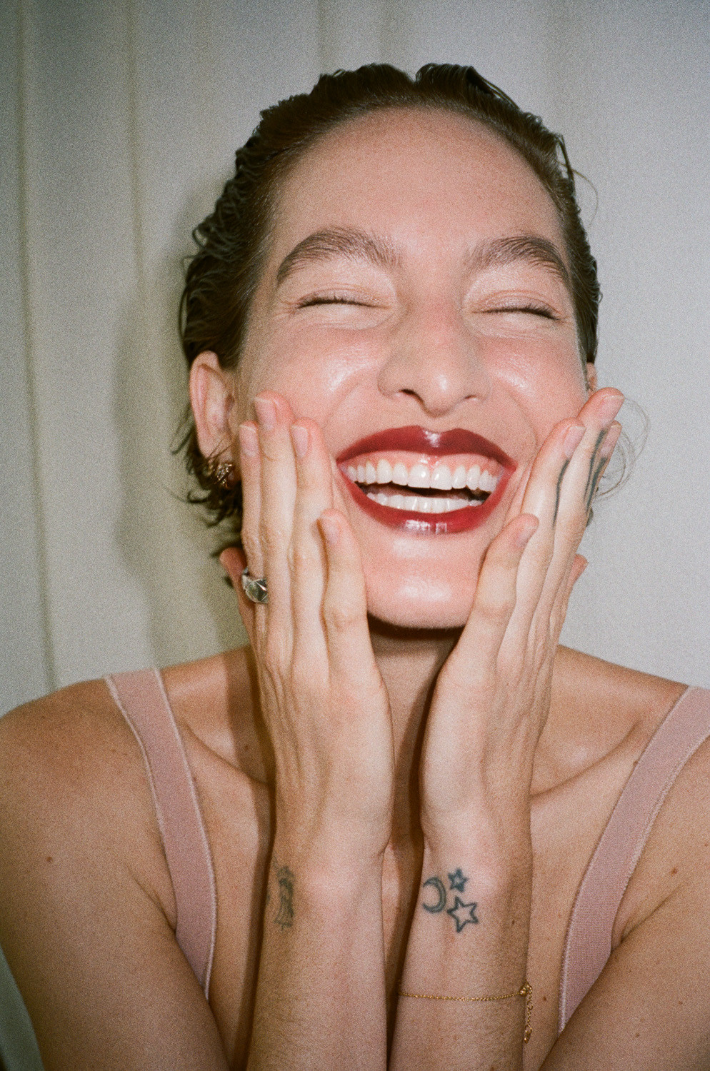 Close-up portrait of a smiling woman with hands on her cheeks, showcasing dark red lip color and tattoos on her wrists, against a light backdrop.
