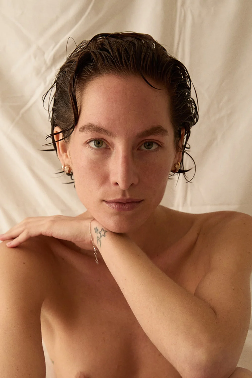 Portrait of a woman with short, wet hair, light skin, and green eyes. She rests her arm on her chest in front of a white sheet.