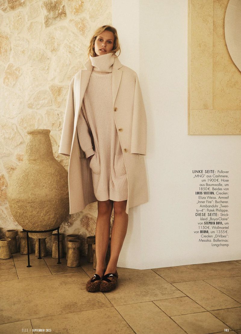 Indoor studio shot of a woman in a sweater dress and coat with stone walls, tile floors, and a large ceramic vase.