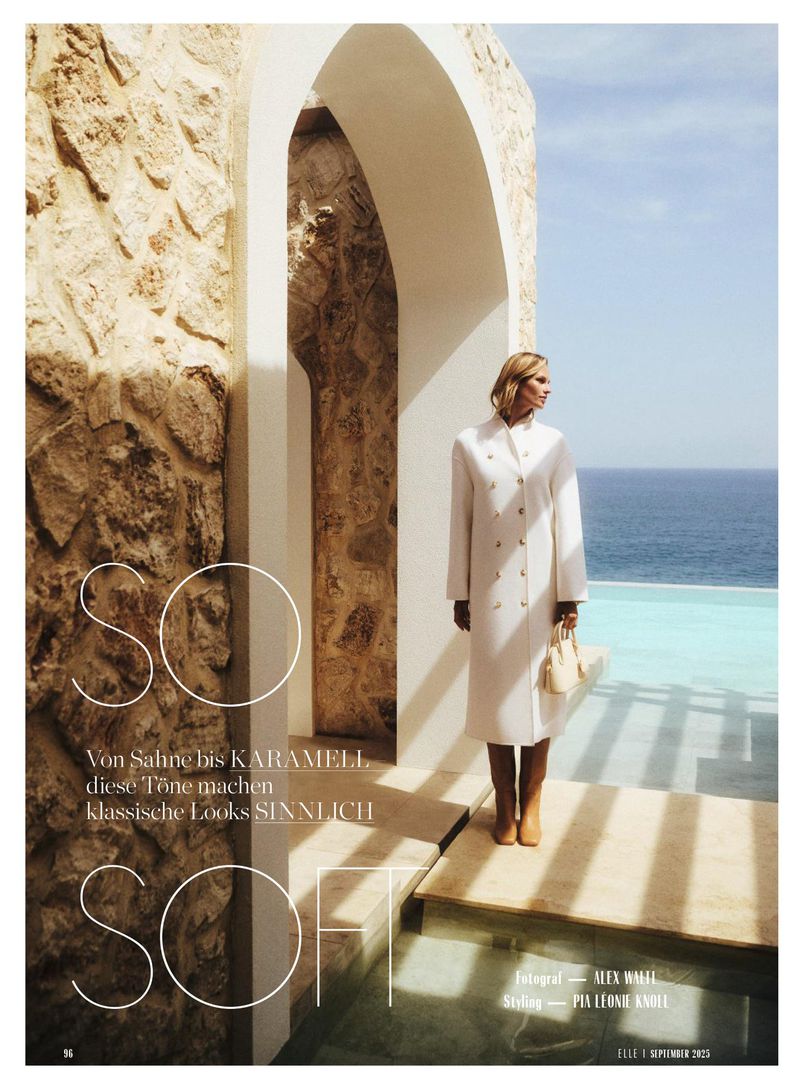 Fashion editorial shot of a blonde woman in a white coat standing near an arched stone wall, with the ocean visible in the background.
