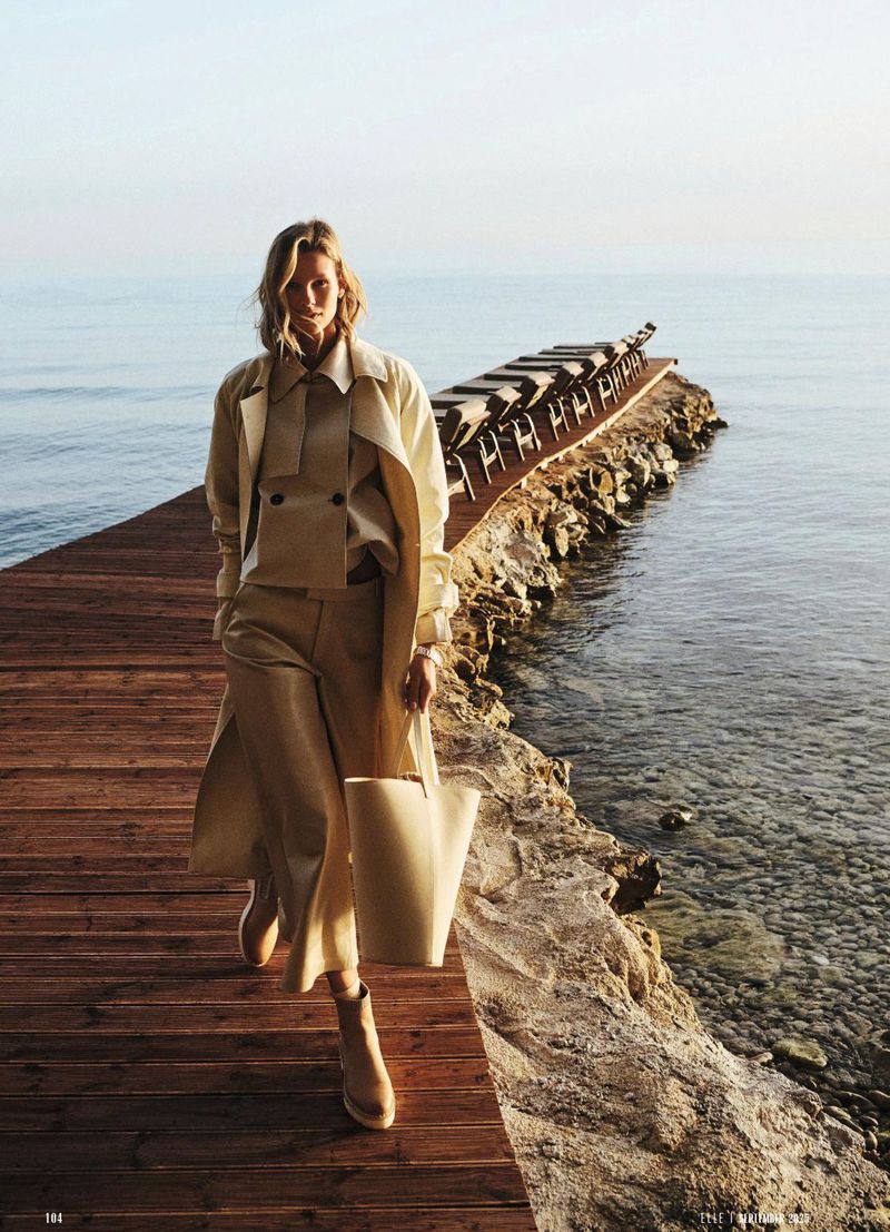 A blonde woman in a beige trench coat and pants walks on a wooden pier near the ocean, carrying a tote bag in a fashion editorial.