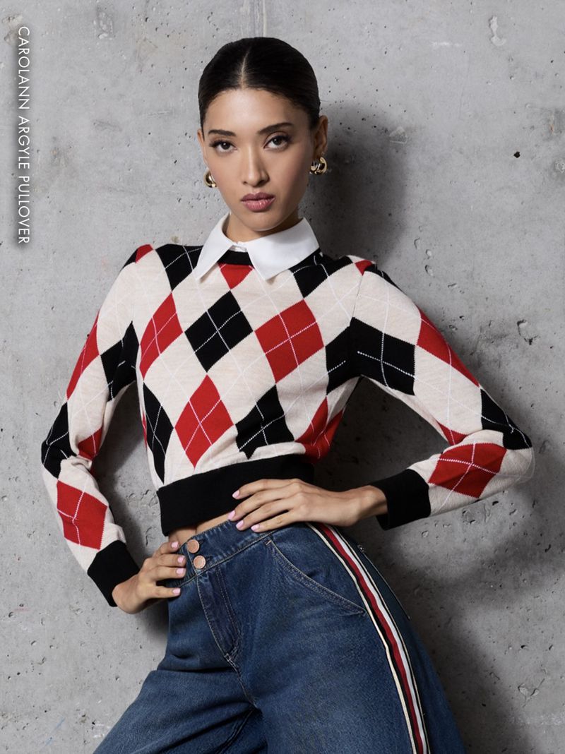 Young woman posing in an argyle sweater with a white collared shirt and wide leg jeans with a red stripe, against a grey background.