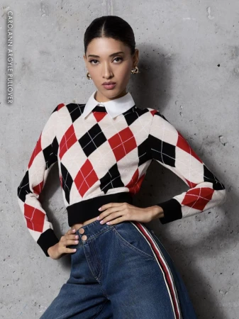 Young woman posing in an argyle sweater with a white collared shirt and wide leg jeans with a red stripe, against a grey background.
