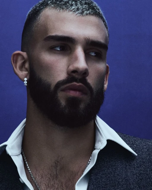 Close-up of a stylish adult male with short grey hair, a beard, and fashionable attire against a plain blue background. He is wearing jewelry.