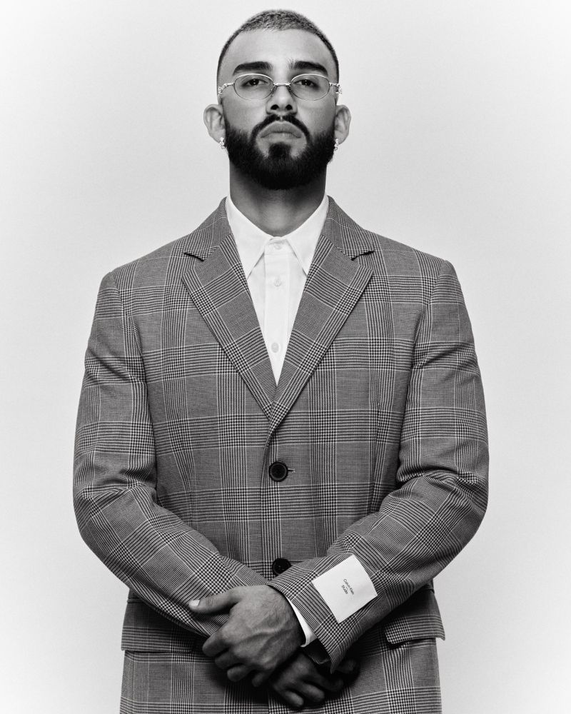 Monochrome portrait of a bearded man in a grey plaid suit with folded arms against a white background in a photo studio setting.
