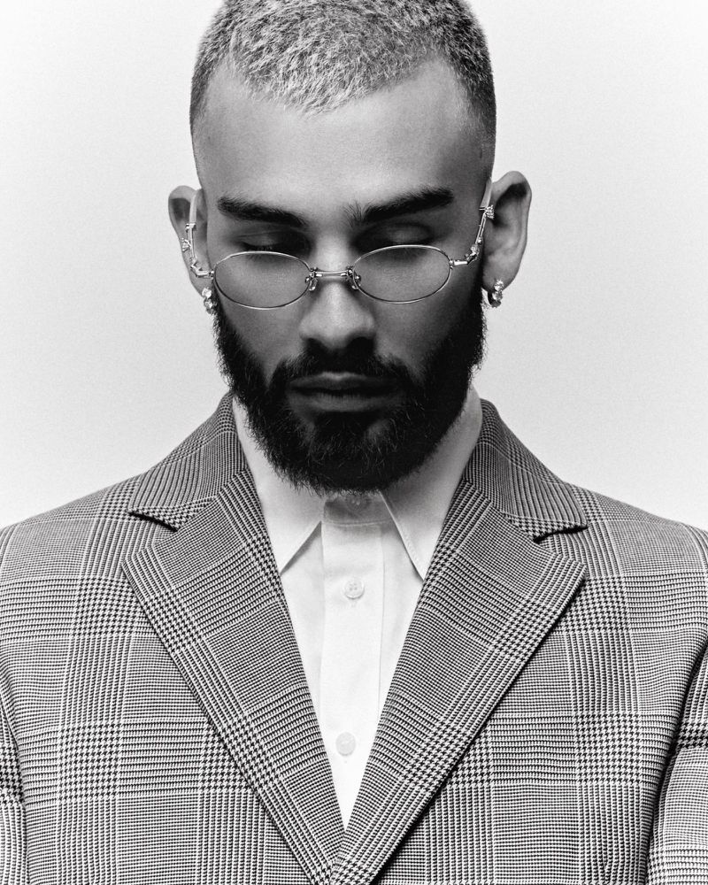 Monochrome portrait of a man with a beard wearing glasses, a white shirt, and a gray plaid suit against a bright white background.