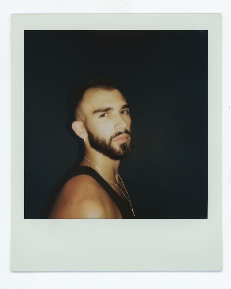 Portrait of a bearded adult male in a black tank top against a dark backdrop, depicted in an instant photo-style frame.