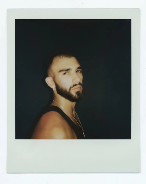 Portrait of a bearded adult male in a black tank top against a dark backdrop, depicted in an instant photo-style frame.