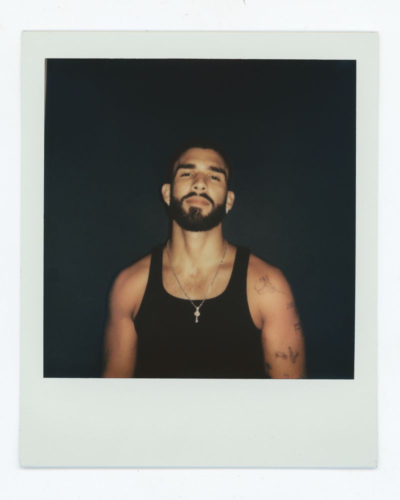 Polaroid portrait of an adult male with a beard wearing a tank top against a dark background, showing shoulders and chest.
